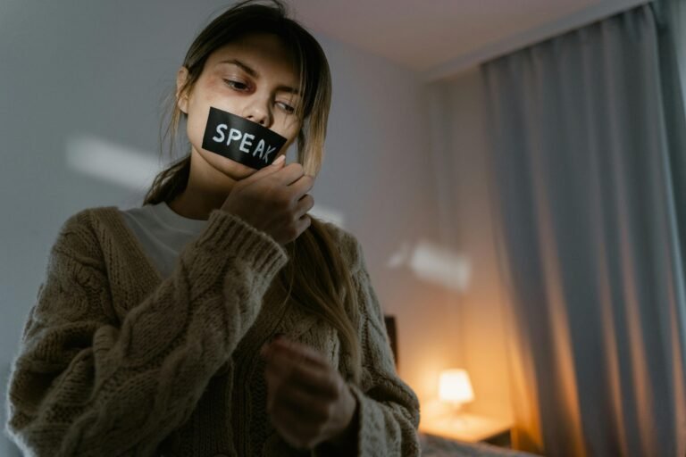 A woman standing in a dimly lit room removing tape with 'SPEAK' written, symbolizing empowerment.